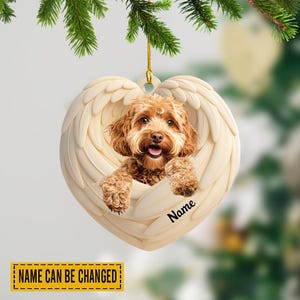 May include: A heart-shaped Christmas ornament featuring a light brown dog nestled within cream-colored angel wings. The dog's name can be added to the ornament. The ornament hangs from a gold string and is surrounded by evergreen branches.