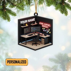 May include: A personalized ornament featuring a miniature computer workstation. The ornament includes a desk, chair, computer monitors, and the text "CODE COFFEE REPEAT." The text "YOUR NAME" is also displayed. The ornament is hanging from a gold string.