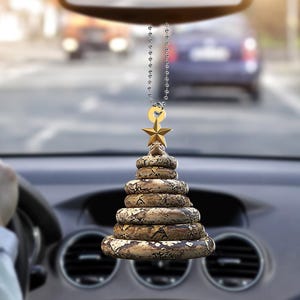 Pu&ograve; includere: Un profumatore per auto a forma di albero di Natale, realizzato con spirali impilate con un motivo a pelle di serpente. Una stella d'oro e un piccolo anello d'oro sono appesi a una catena d'argento. Il profumatore &egrave; appeso allo specchietto retrovisore di un'auto.