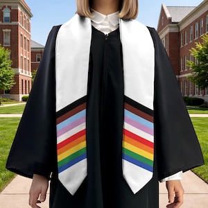 May include: A black graduation gown with a white sash featuring a rainbow and brown stripe design. The sash is draped over the shoulders, with the colorful stripes visible. The gown is worn by a person in front of a brick building.