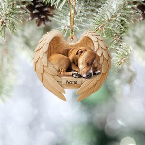 May include: A heart-shaped ornament featuring a sleeping dog nestled within angel wings. The dog is brown with white markings, and the wings are a light beige color. A small plaque at the base reads "Name". A gold string is attached for hanging.