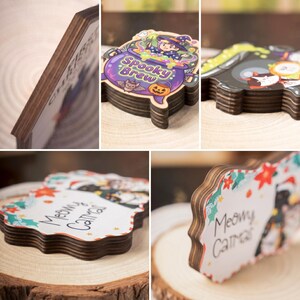 May include: Wooden coasters with decorative designs. One coaster features a witch in a cauldron with the text "Spooky Brew". Another coaster has the text "Meowy Catmas" with a cat illustration. The coasters have a dark brown edge.