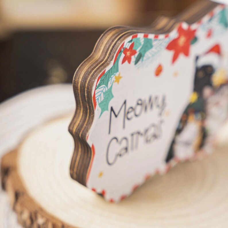 May include: A wooden sign with a scalloped edge, featuring a Christmas-themed design. The sign has a white background with a festive border of red poinsettias, green holly, and stars. The text "Meowy Christmas" is handwritten in black script, with images of cats.