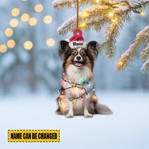 May include: A Christmas ornament featuring a Papillon dog wearing a Santa hat with the word "Name" and a string of colorful lights. The text "NAME CAN BE CHANGED" is at the bottom. The background is a snowy winter scene.