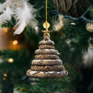 Pu&ograve; includere: Un originale ornamento natalizio a forma di albero fatto di serpenti impilati. I serpenti hanno un motivo marrone e beige. Una stella d'oro &egrave; in cima, e un anello d'oro &egrave; in alto per appenderlo. L'ornamento &egrave; appeso davanti a un albero di Natale sfocato.
