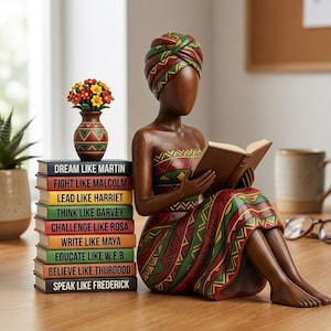 May include: A brown figurine of a woman reading a book, wearing a patterned dress and head wrap. A stack of books with inspirational phrases like "Dream Like Martin" and a vase with flowers are next to her. The figurine is on a wooden surface.