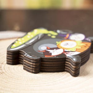 May include: A stack of wooden coasters with a cartoon graphic. The top coaster features a black background with a cartoon character design, including green, orange, and white elements. The coasters have a dark brown, layered edge.