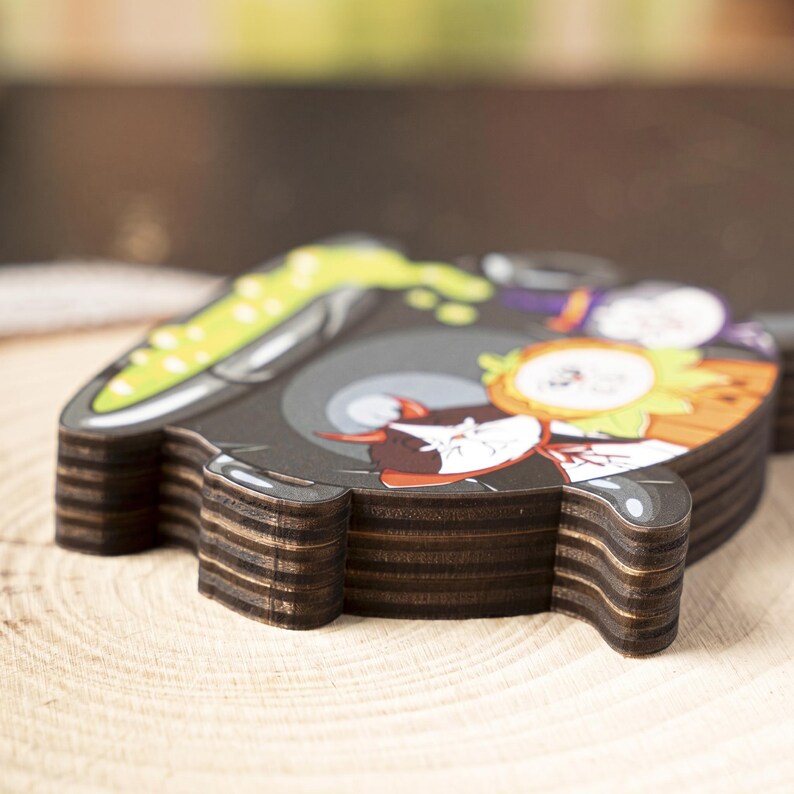 May include: A stack of wooden coasters with a cartoon graphic. The top coaster features a black background with a cartoon character design, including green, orange, and white elements. The coasters have a dark brown, layered edge.