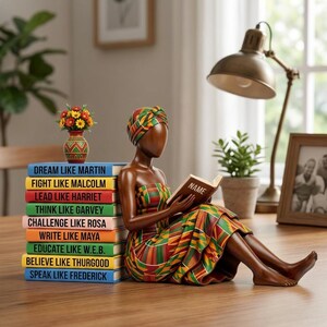 May include: A wooden figurine of a woman in colorful African attire, reading a book with the word "NAME" on the cover. A stack of books with inspirational phrases like "Dream Like Martin" and "Fight Like Malcolm" are next to her. A small vase with flowers sits atop the books.