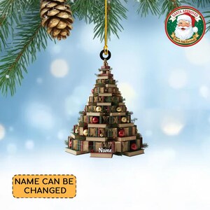 Puede incluir: Adorno de árbol de Navidad con forma de árbol, construido con libros apilados, adornado con bolas rojas y doradas. El adorno tiene un lazo dorado para colgar y el texto "Name" es visible. Las palabras "Name Can Be Changed" están en un rectángulo amarillo. Un sello "Santa Approved" está en la parte superior derecha.