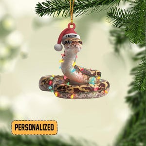 May include: A Christmas ornament featuring a snake wearing a Santa hat and string lights. The ornament has a gold string for hanging and the word "Name" is printed on the hat. The word "PERSONALIZED" is printed on a yellow rectangle.