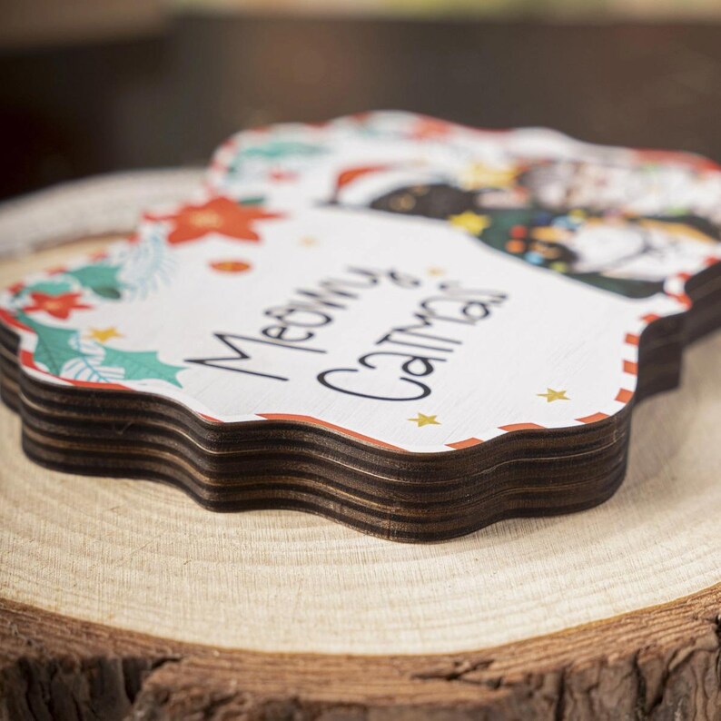 May include: A stack of wooden coasters with a Christmas theme. The coasters are white with a decorative border and the words "Meowy Christmas" in black script. The coasters have a cat wearing a Santa hat and are decorated with holly, stars, and poinsettias.