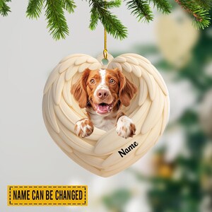May include: Heart-shaped ornament featuring a dog with brown and white fur, framed by cream-colored angel wings. The dog is smiling with its tongue out. The word "Name" is written on the ornament. The text "NAME CAN BE CHANGED" is on a yellow banner.
