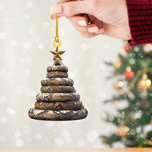 Pu&ograve; includere: Un ornamento per l'albero di Natale a forma di albero, realizzato con anelli impilati e strutturati con un motivo a serpente. Una stella d'oro &egrave; in cima, con un anello e un cordino dorati per appenderlo. L'ornamento &egrave; tenuto da una mano.