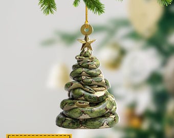 Può includere: Un ornamento per l'albero di Natale a forma di albero fatto di serpenti verdi e beige. Ha una stella d'oro e un anello d'oro per appenderlo. Il testo "NAME CAN BE CHANGED" è su un rettangolo giallo.
