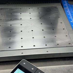 Puede incluir: Una placa metálica plateada con una cuadrícula de pequeños agujeros y líneas grabadas. El texto "F2 Ultra" es visible. Una esterilla de corte azul y un pequeño dispositivo electrónico están en el fondo.