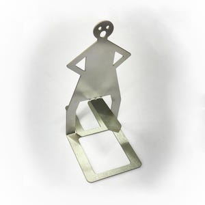 May include: A silver-coloured metal bookend shaped like a stick figure with arms akimbo. The figure has a surprised expression and stands on a rectangular base. The bookend is designed to hold books upright.