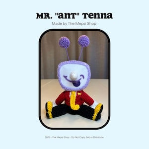 May include: A crocheted doll in the shape of a television with the text "MR. 'ant' tenna" and "Made by The Mepsi Shop". The doll has a red jacket, black pants, and yellow accents. The TV screen has a smiling face with a tongue sticking out.
