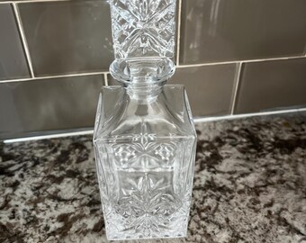 May include: A clear, square-shaped glass decanter with a matching stopper. The decanter and stopper feature a cut-glass design with starburst and geometric patterns. The stopper is rectangular and fits snugly into the decanter's neck. The decanter is likely used for storing and serving spirits.
