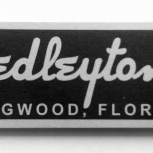 Puede incluir: Una placa de metal plateado con fondo negro. El texto "Hedleytone" est&aacute; escrito en una fuente blanca cursiva. Debajo del texto se encuentra la ubicaci&oacute;n "Longwood, Florida".
