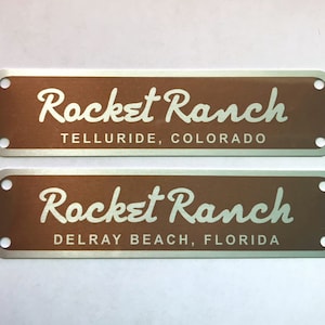 Puede incluir: Dos placas rectangulares de metal con esquinas redondeadas. La placa superior es marr&oacute;n con texto blanco que dice "Rocket Ranch Telluride, Colorado". La placa inferior es marr&oacute;n con texto blanco que dice "Rocket Ranch Delray Beach, Florida".