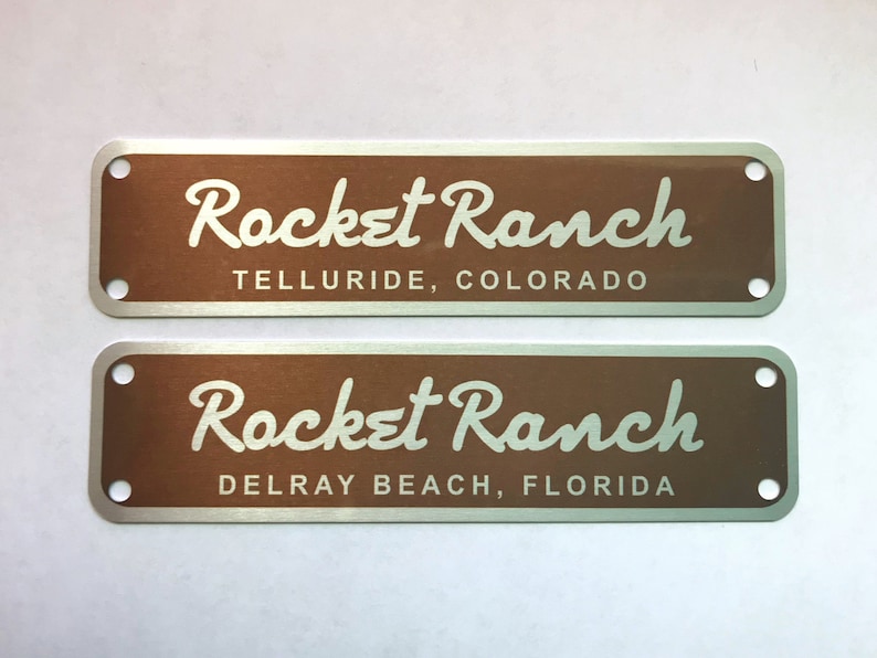 Puede incluir: Dos placas rectangulares de metal con esquinas redondeadas. La placa superior es marr&oacute;n con texto blanco que dice "Rocket Ranch Telluride, Colorado". La placa inferior es marr&oacute;n con texto blanco que dice "Rocket Ranch Delray Beach, Florida".