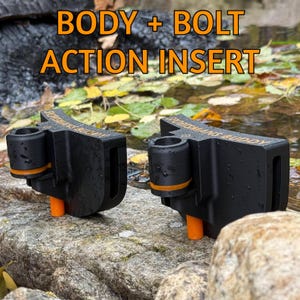 May include: Two black "Body + Bolt Action Insert" devices with orange accents and pegs, resting on a rock. The text "BODY + BOLT ACTION INSERT" is in orange above the devices. The devices are near water with leaves.
