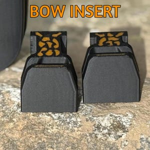 May include: Two black bow inserts with a decorative gold and black pattern on top. The text "BOW INSERT" is displayed in orange at the top of the image. The inserts are sitting on a textured, light brown surface.