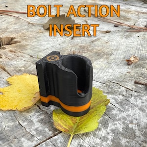 May include: Black and orange bolt action insert. The insert has a curved design with an orange stripe and the text "BOLT ACTION INSERT" in orange. The insert is resting on a wooden surface with yellow and green leaves.