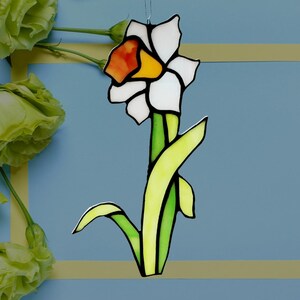 May include: A stained glass daffodil with white petals, a yellow and orange center, and green stem and leaves. The flower is outlined in black and hangs against a blue background.