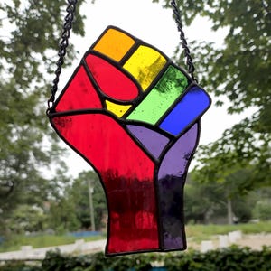 May include: A stained glass fist in a rainbow of colors, including red, orange, yellow, green, blue, and purple. The fist is outlined in black and hangs from a black chain. The background is blurred with green foliage.