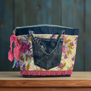 May include: A handmade tote bag with a denim pocket and trim. The bag features a floral pattern with pink, purple, and green flowers on a light background. A pink lace trim and a zipper closure complete the design.