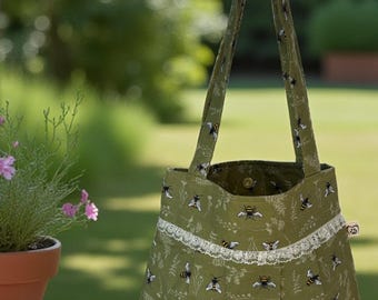 May include: A green tote bag with a bee and floral pattern. The bag has a white lace trim and matching handles. The bag is sitting on a wooden surface with a potted plant in the background.