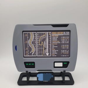 May include: A grey rectangular display screen mounted on a black stand, featuring a medical diagram titled HUMAN ANATOMY-03.7329. The screen shows a yellow and white illustration of a human spine and vertebrae with detailed text labels and data.