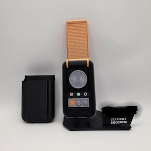 Może przedstawiać: Czarny rekwizyt Starfleet Tricorder z pomarańczową teksturowaną pokrywą, okrągłym szarym czujnikiem, trzema małymi kolorowymi przyciskami i prostokątnym panelem sterowania na czarnej podstawce z napisem STARFLEET TRICORDER.