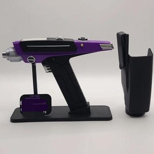 May include: A purple and black Star Trek phaser prop replica mounted on a black display stand with a small rectangular plaque featuring the text Star Trek. A matching black holster is positioned to the right of the phaser on the stand.