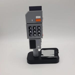 May include: A gray, vertical, block-shaped device with a numeric keypad featuring black buttons with white numbers one through nine. It includes a small orange button, a black top section, and a textured gray panel, all mounted on a black base.