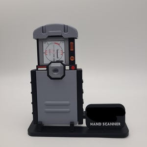 Puede incluir: Un escáner manual gris y negro con una pantalla rectangular que muestra un gráfico de medidor impreso, una lente circular central y una base negra con un pequeño letrero que dice HAND SCANNER en letras blancas mayúsculas.