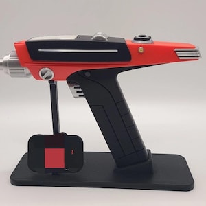 May include: A sci-fi style prop phaser weapon featuring a bright orange upper body with black accents, a silver metallic nozzle, and a black handle. The device is mounted on a black rectangular display stand with a vertical support arm.
