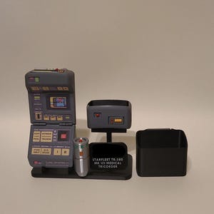May include: A grey Starfleet TR-580 MK VII medical tricorder prop set on a black display stand. The device features a hinged screen with various labels, a small silver handheld scanner, and a separate black storage container component.