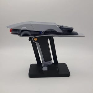 May include: A gray and black prop phaser replica mounted on a black display stand. The device features a long, tapered top section, a textured black handle with a gold circular accent, and a red detail at the rear. The stand has a rectangular base.