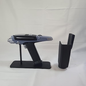 May include: A replica phaser prop with a black handle and a gray body, displayed on a black stand. The prop includes a separate black holster. The design is reminiscent of a science fiction weapon.