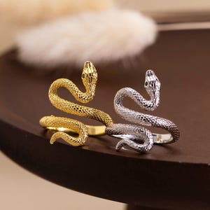 May include: Two snake-shaped rings, one in a gold-tone finish and one in a silver-tone finish. Each ring features a textured, scale-like surface pattern that wraps around the finger, with the head of the snake raised at the top.