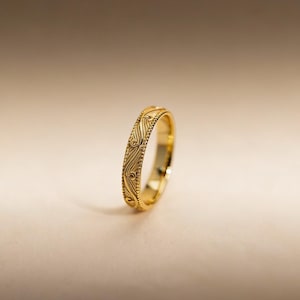 May include: A gold ring featuring a polished finish and a central band decorated with an intricate, repeating engraved swirl pattern, bordered on both sides by a delicate beaded edge detail that runs along the entire circumference.