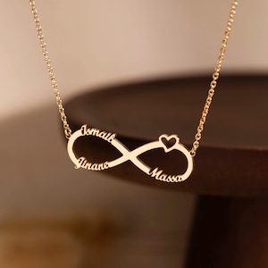 Op de afbeelding: Een goudkleurige metalen ketting met een hanger in de vorm van een oneindigheidssymbool. De hanger is geïntegreerd in een fijne ketting en bevat de namen Ismail, Yinane en Massa gegraveerd langs de lussen, met een klein hartje rechtsboven.