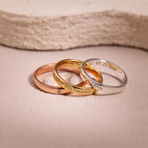 May include: Three rings in rose gold, gold, and silver. The gold ring has "I love you Forever" engraved. The silver ring has "07.22.2024" and "Oliver" engraved. The rose gold ring has a letter "R" engraved. The rings are on a pink surface.