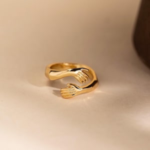 Peut inclure: Une bague dorée représentant deux mains enlacées. La bague est en métal poli et se détache sur un fond neutre. Les mains sont détaillées, avec des doigts visibles et une finition lisse. Le design de la bague évoque des thèmes d'amour et de connexion.