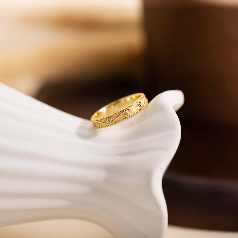 May include: A gold-colored ring with an intricate, repeating pattern. The ring is set against a white, curved surface, with a blurred background of brown and beige tones. The ring's design features a series of small, detailed shapes.