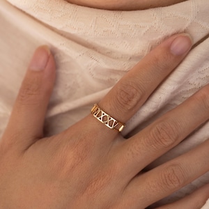 Può includere: Un anello color oro con numeri romani MXXV è mostrato su un dito. L'anello ha un design aperto, con i numeri ritagliati. Lo sfondo è di un colore morbido e neutro.