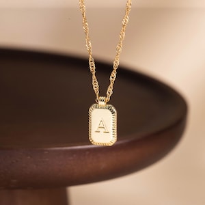 May include: A gold-colored necklace with a rectangular pendant. The pendant features the letter "A" in the center. The chain is a twisted rope design. The necklace is displayed against a blurred, dark brown background.